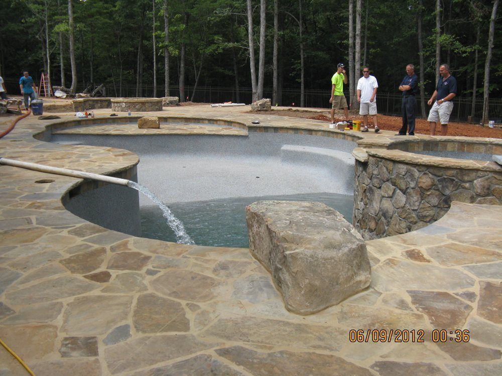 Stone patio surrounding a circular fountain basin at a park, with people standing nearby and trees in back