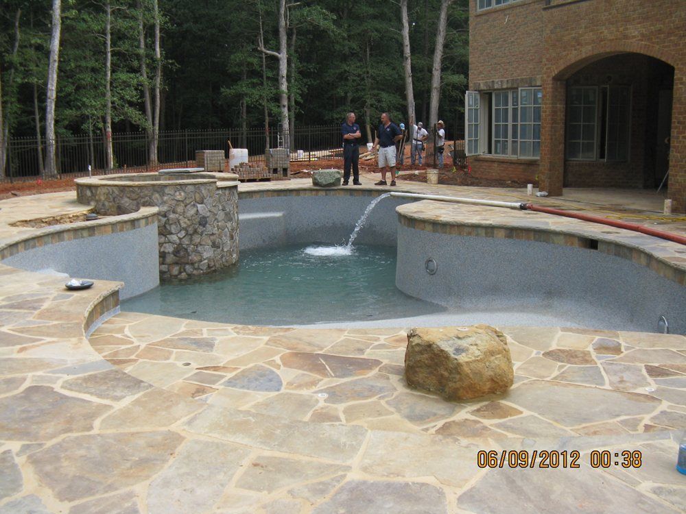 Stone patio with a circular pool and fountain beside a building and trees, with a few people in the background