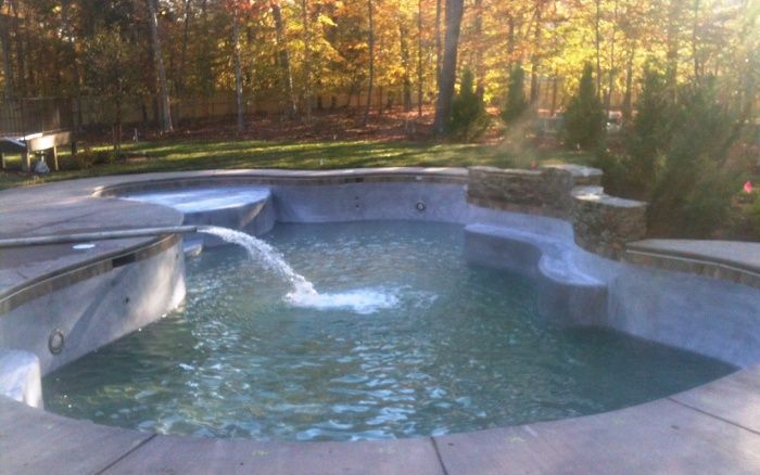 Empty backyard pool with a built-in hot tub and water jet spraying, surrounded by autumn trees