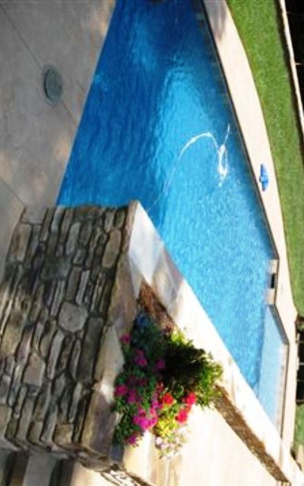 Blue swimming pool beside stone wall and white walkway with pink flowers and green lawn