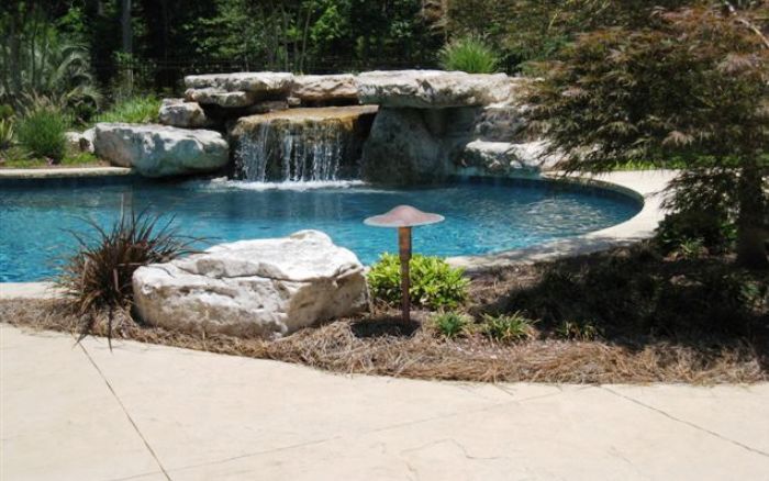 Backyard pool with waterfall, large rocks, and surrounding greenery beside a paved patio