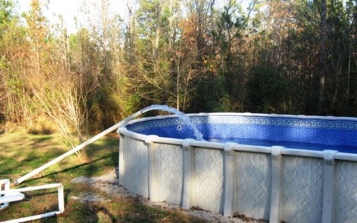 An above-ground pool with a white hose and blue cover in a wooded backyard