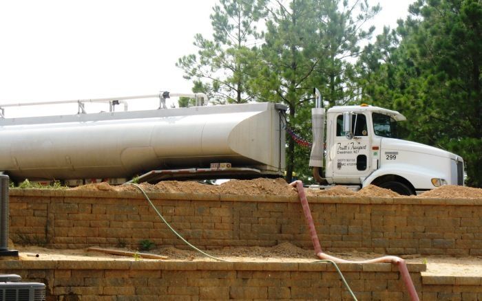 White tanker truck parked beside a brick wall with hoses on the ground outdoors