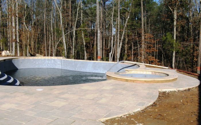 Empty backyard pool with a circular hot tub, stone deck, and wooded trees in the background