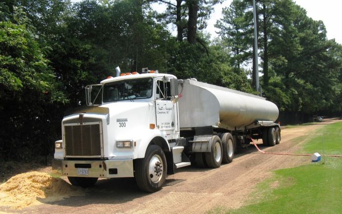 White tanker truck parked on a dirt road beside a pile of tan spill material in a wooded area