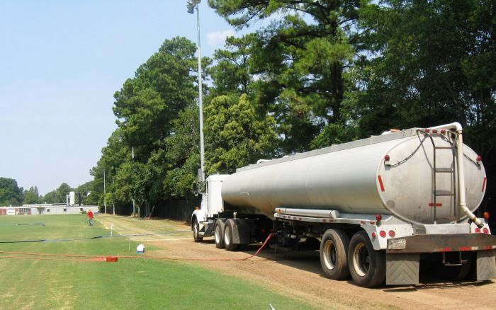 White tanker truck parked on a grassy roadside beside trees and a field