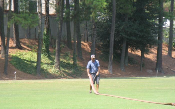 Person standing on a grassy field holding a long rope near trees in the background