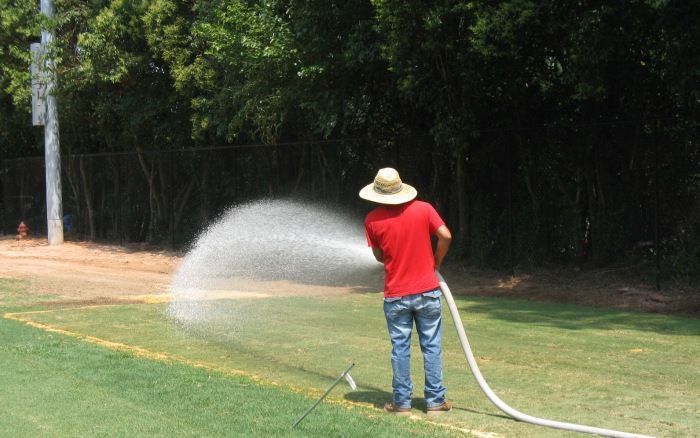 Person in a red shirt watering a grassy field with a hose under trees