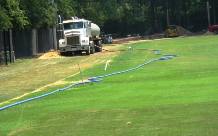 Water truck spraying a long green lawn beside a tree-lined road, with blue hoses laid across the grass