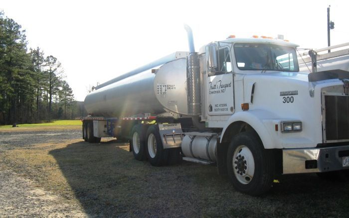 White tanker truck parked on gravel beside trees on a sunny day
