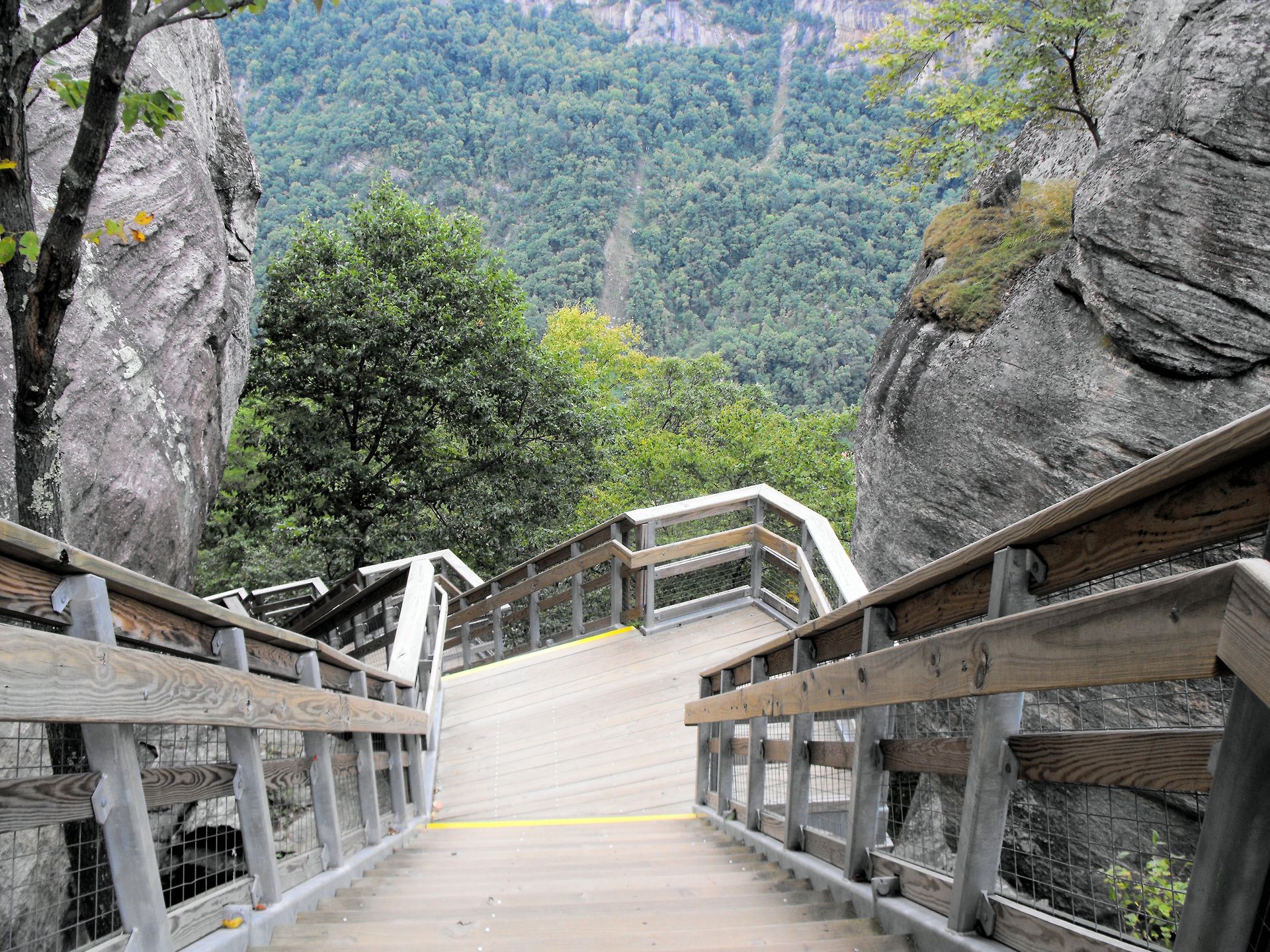 A wooden staircase with railings descends between two large rock faces, looking out over a forested mountain valley. A wooden staircase with railings descends between two large rock faces, looking out over a forested mountain valley.