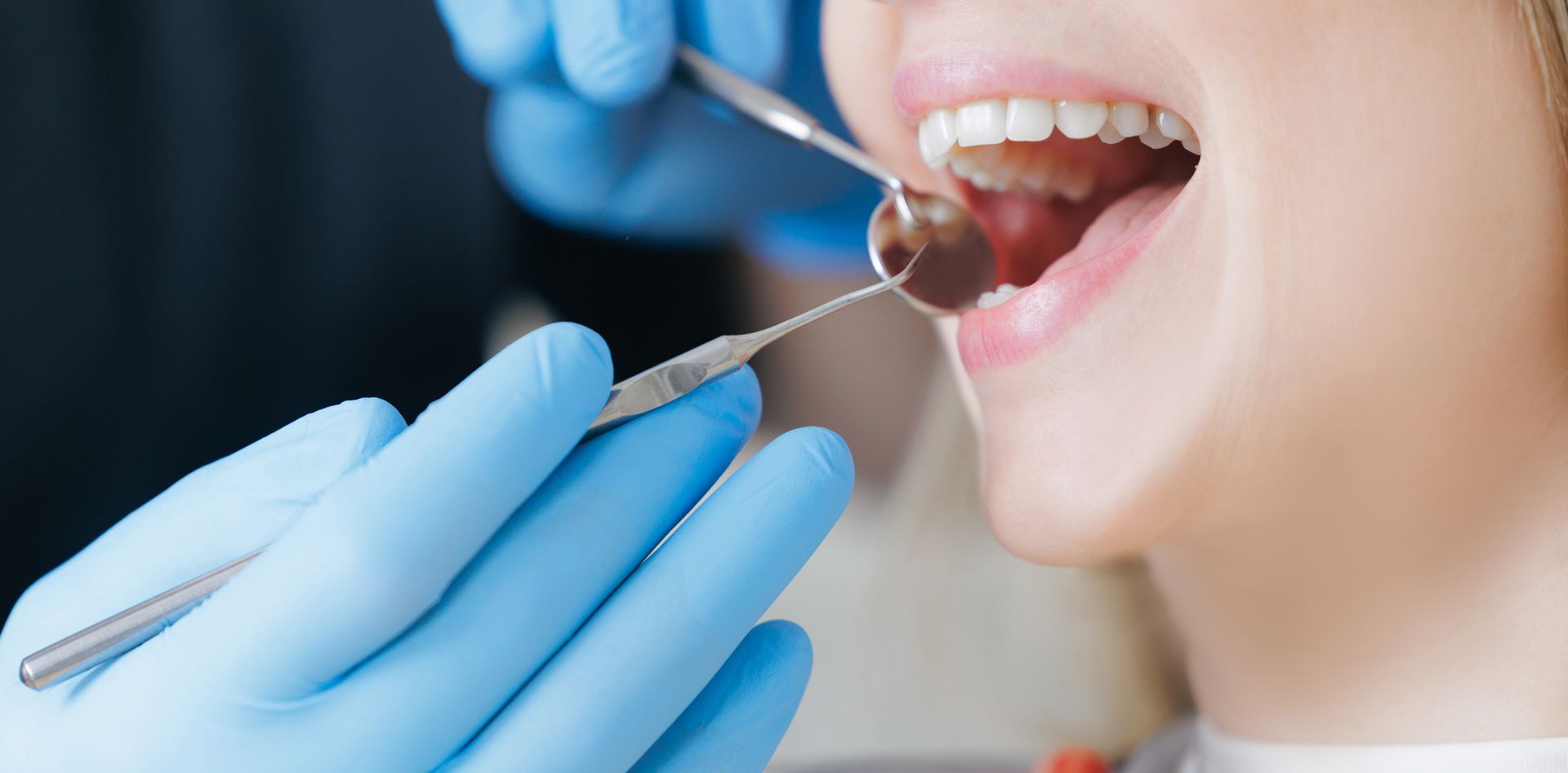 A dentist wearing blue gloves uses a dental mirror to examine a patient's teeth. A dentist wearing blue gloves uses a dental mirror to examine a patient's teeth.
