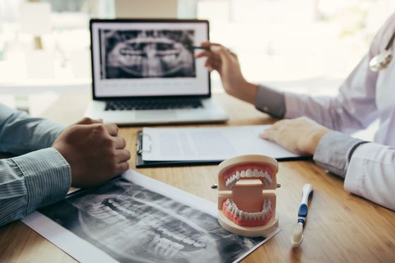 A dentist consults with a patient, reviewing a dental X-ray on a laptop next to a model of teeth and a toothbrush. A dentist consults with a patient, reviewing a dental X-ray on a laptop next to a model of teeth and a toothbrush.