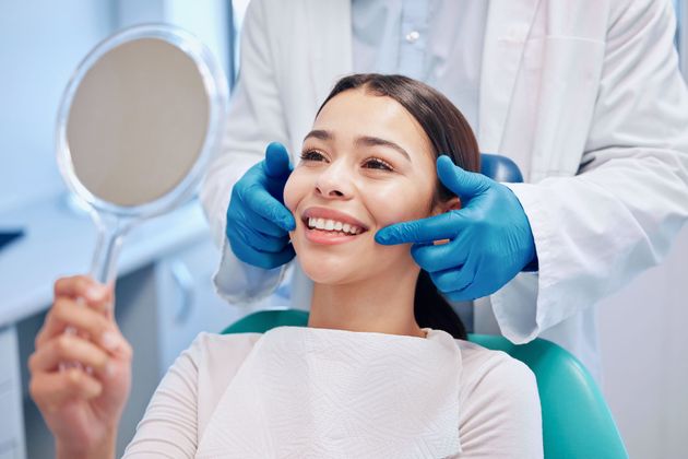 A smiling patient in a dental chair looks into a handheld mirror while a dentist in blue gloves adjusts her cheeks. A smiling patient in a dental chair looks into a handheld mirror while a dentist in blue gloves adjusts her cheeks.
