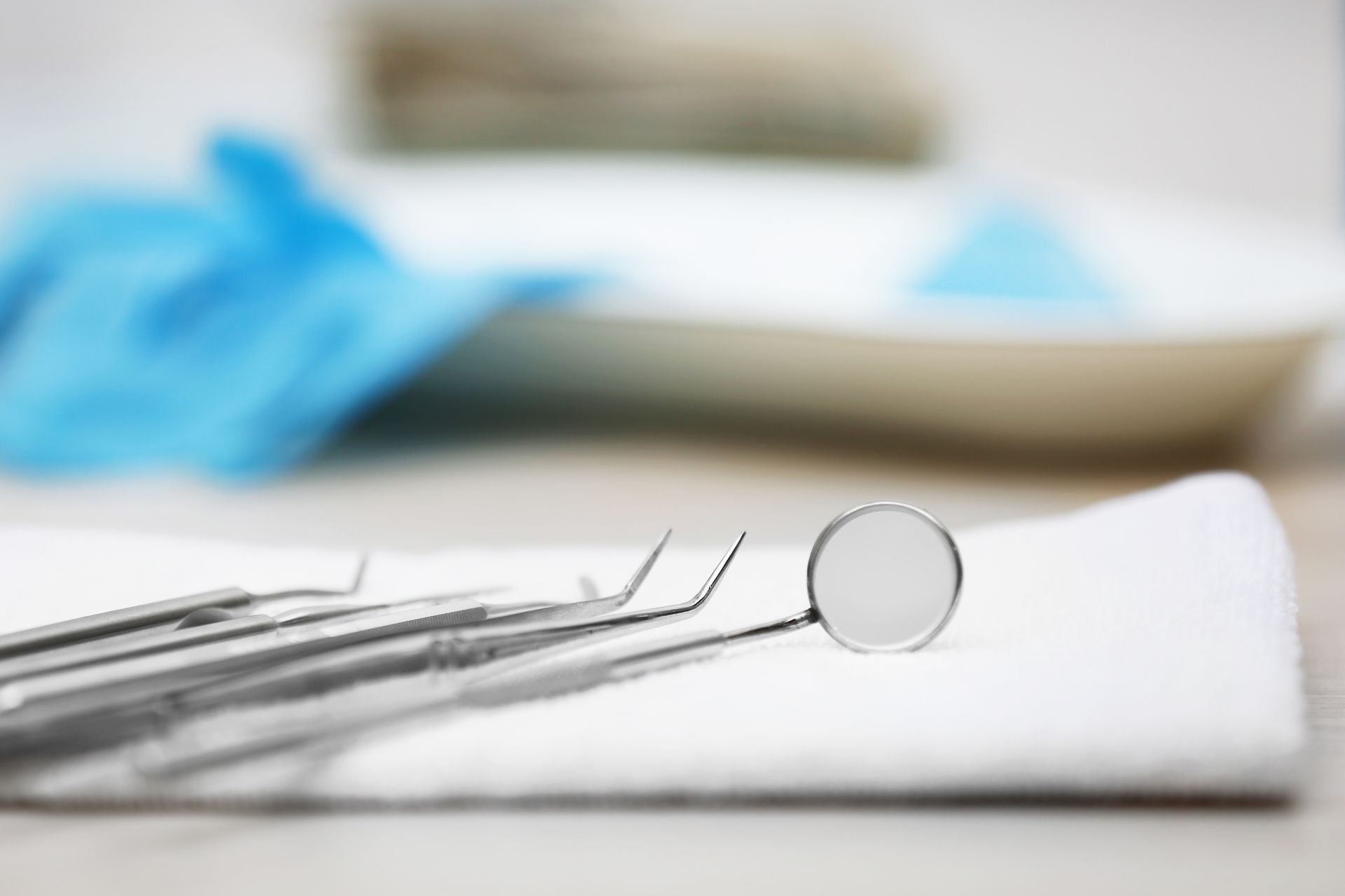 A dental mirror and metal tools rest on a white cloth, with blue medical gloves blurred in the background. A dental mirror and metal tools rest on a white cloth, with blue medical gloves blurred in the background.