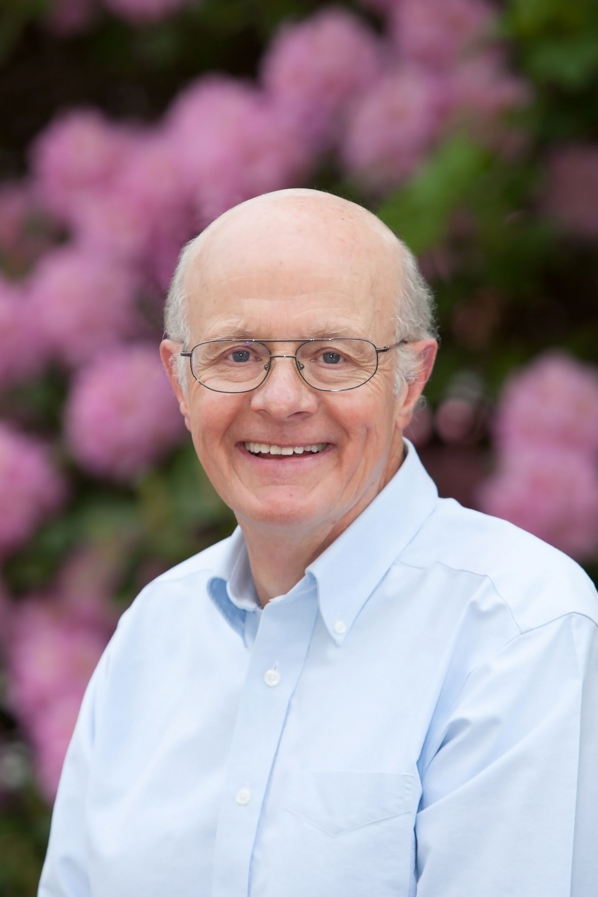 A person with glasses and a light blue button-down shirt smiling in front of a background of soft pink flowers. A person with glasses and a light blue button-down shirt smiling in front of a background of soft pink flowers.