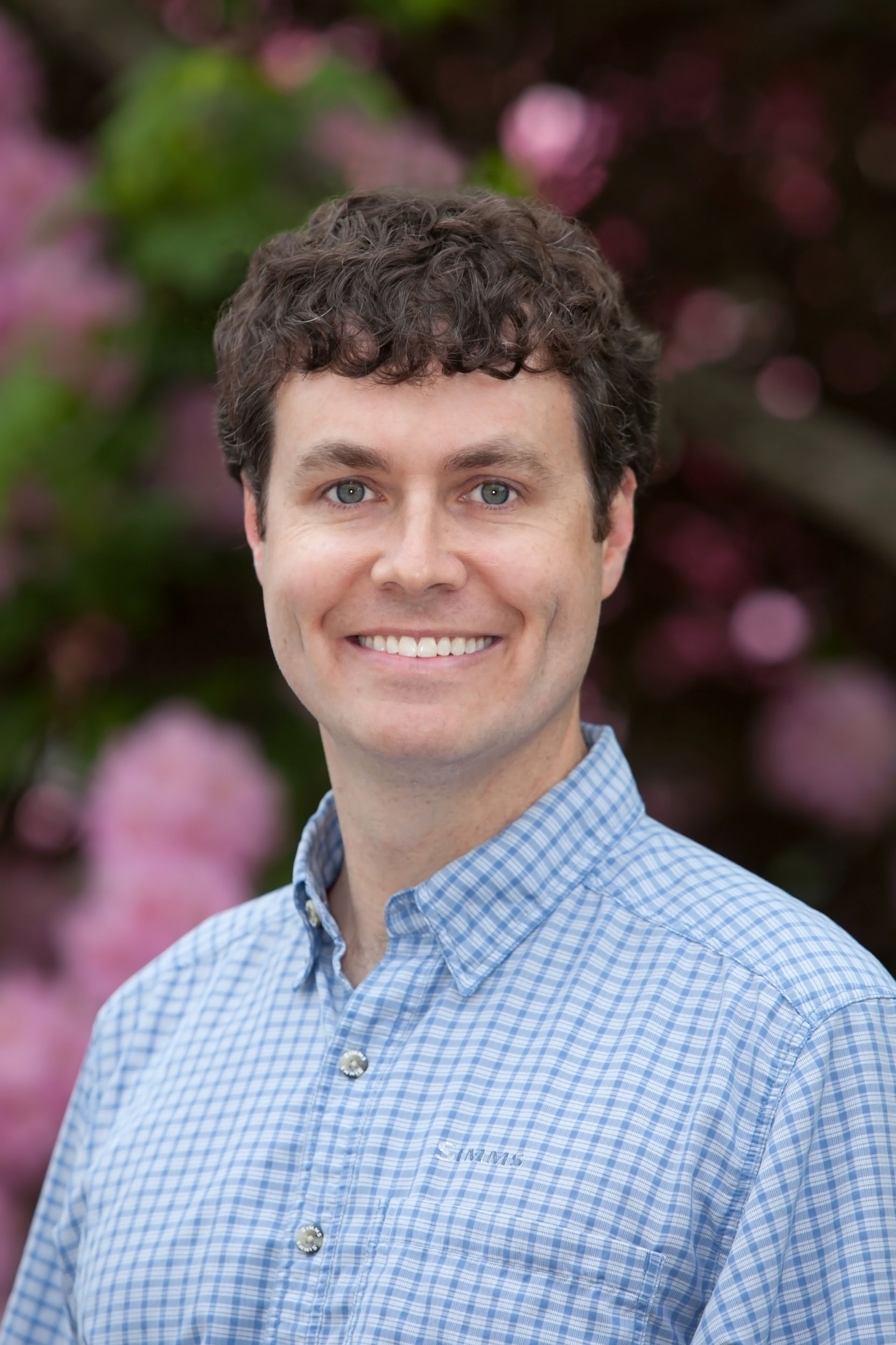 A person with curly hair smiles while wearing a light blue checkered shirt, set against a blurred, floral background. A person with curly hair smiles while wearing a light blue checkered shirt, set against a blurred, floral background.