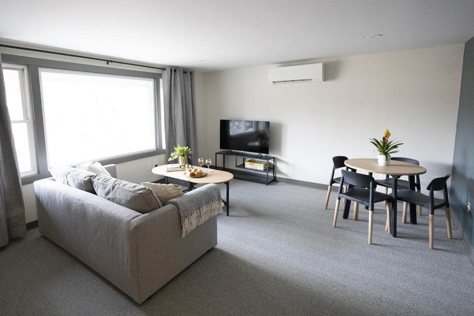 Modern living room with gray sofa, TV, coffee table, and small dining table by a bright window.