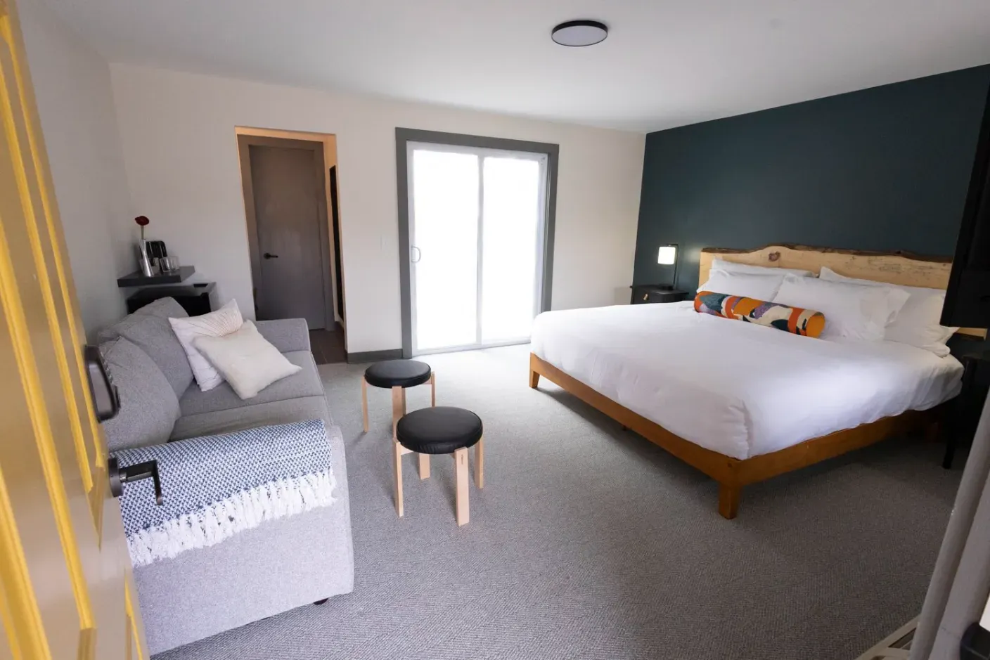 Modern bedroom with a large bed, sofa, small stools, and a sliding glass door.