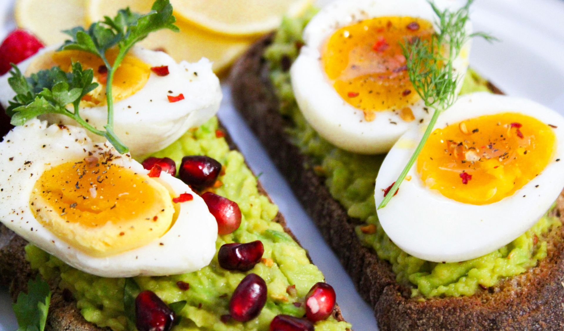 Avocado toast topped with sliced boiled eggs, pomegranate seeds, and herbs on a white plate