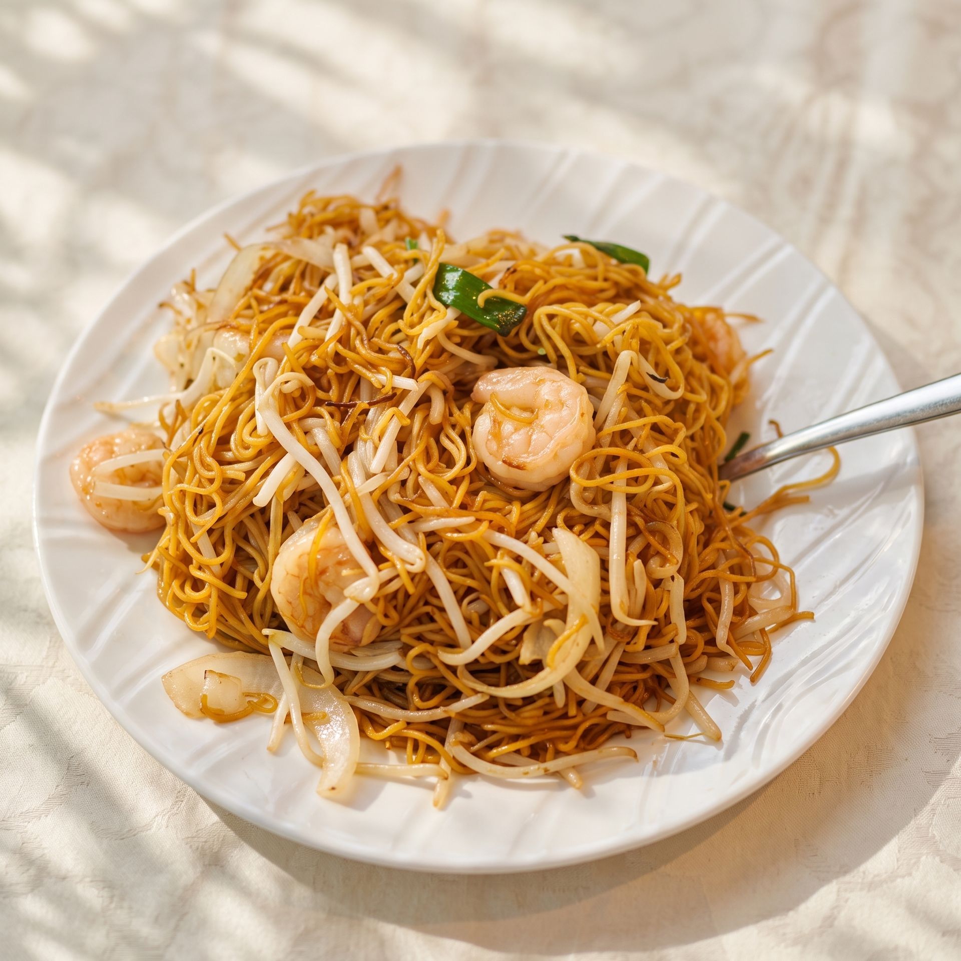 Plate of shrimp chow mein noodles with bean sprouts, served on a white dish with a fork on a light table.