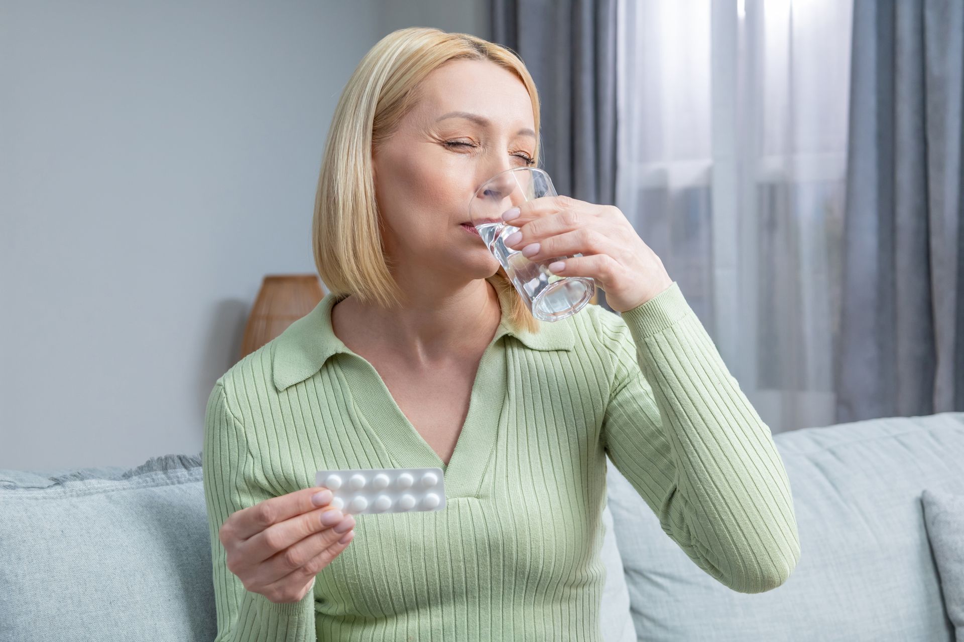 A blonde woman in her fifties drinks a glass of water while holding a packet of pills, inside a room. A blonde woman in her fifties drinks a glass of water while holding a packet of pills, inside a room.