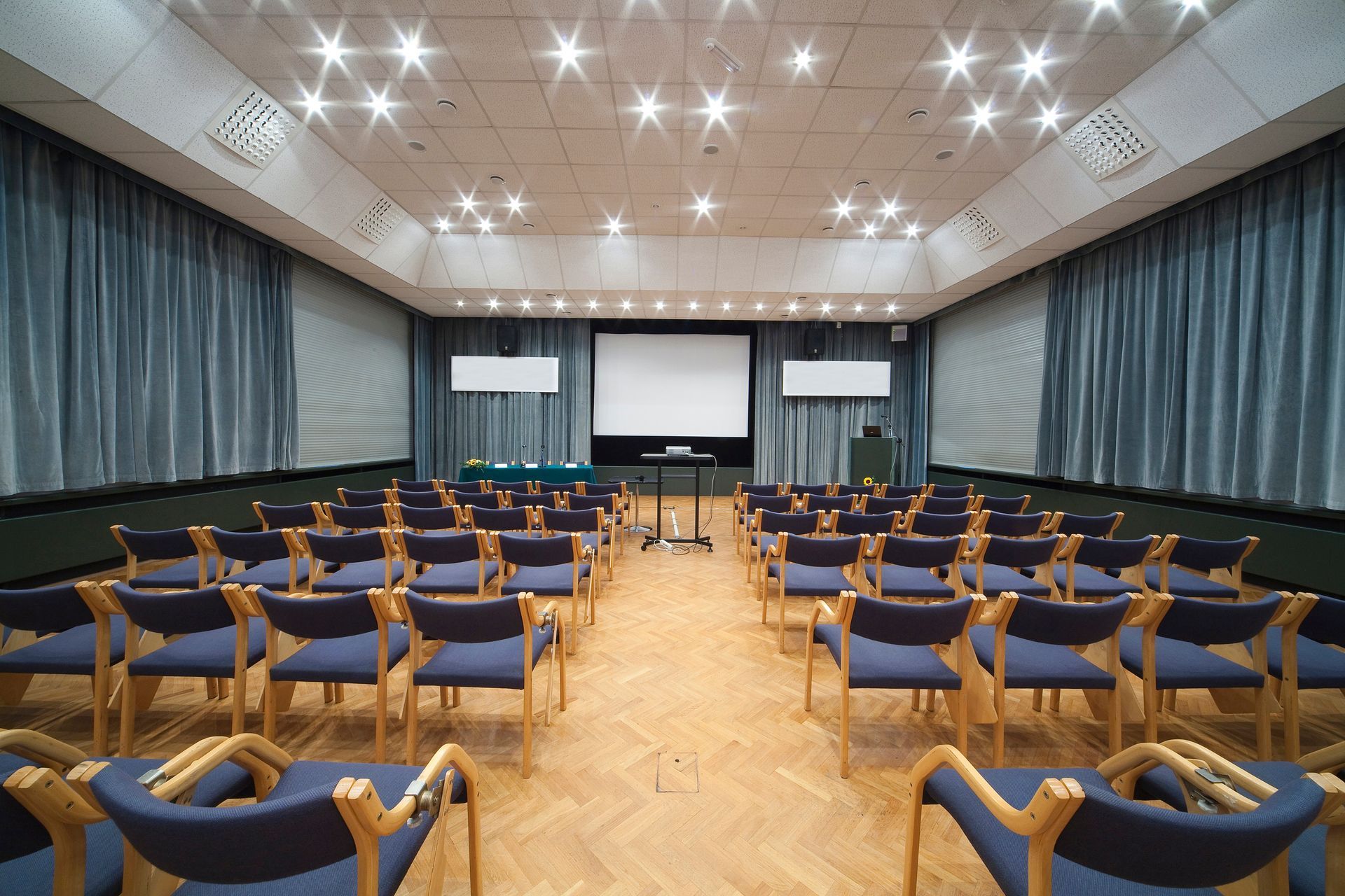 Empty conference room with rows of blue chairs, wood floor, and a projector screen at the front