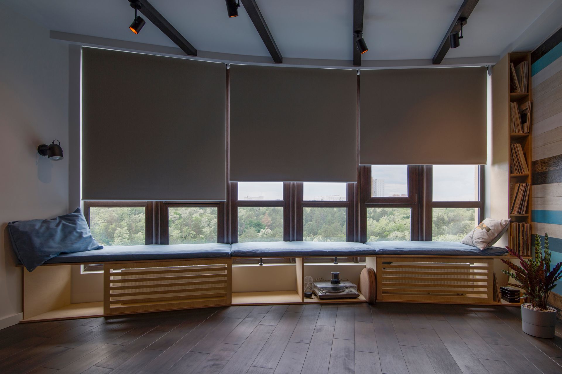 Modern living room with large windows, gray blinds, wooden benches, and a potted plant on the right