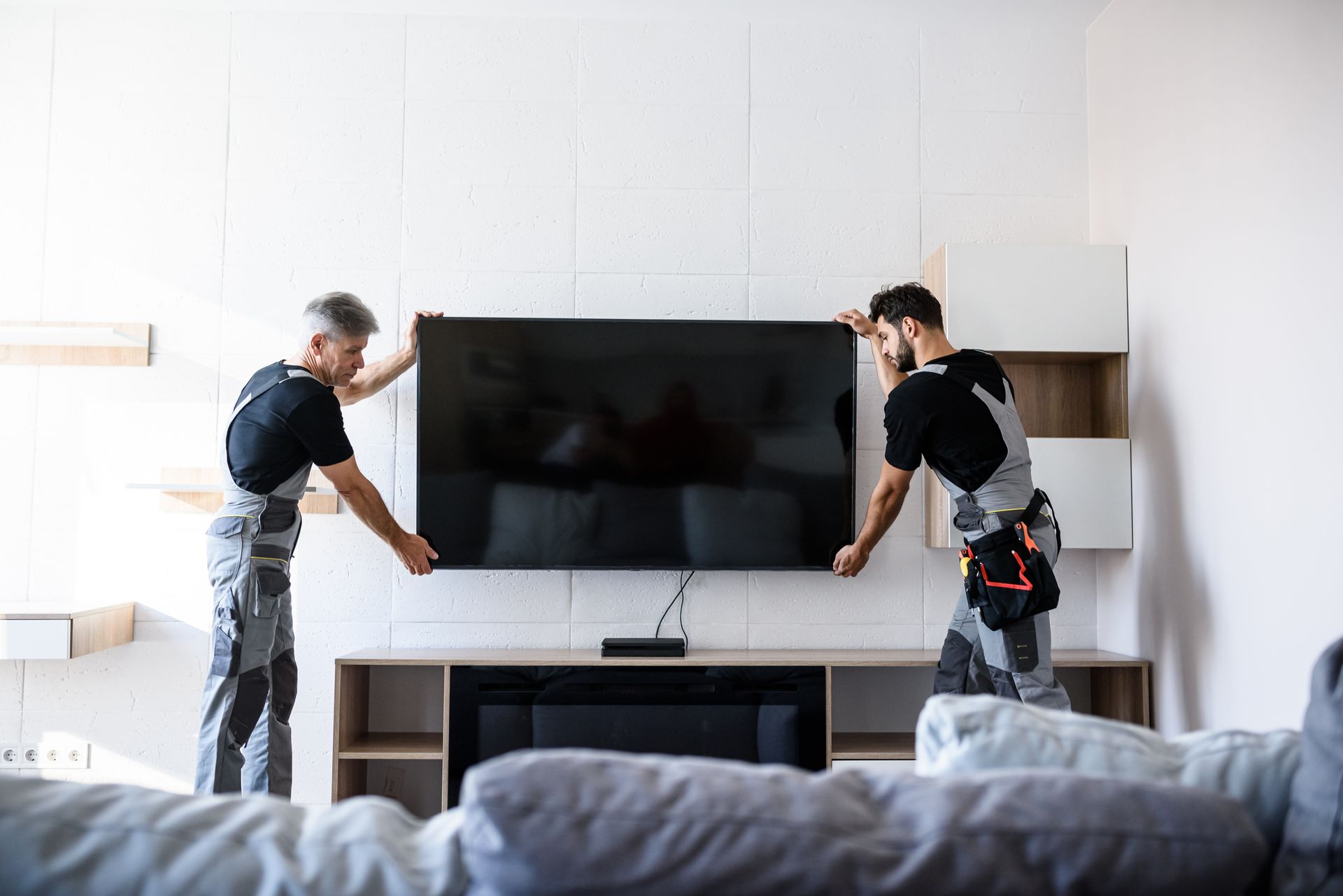 Two people mounting a large TV on a wall above a media console in a bright living room.