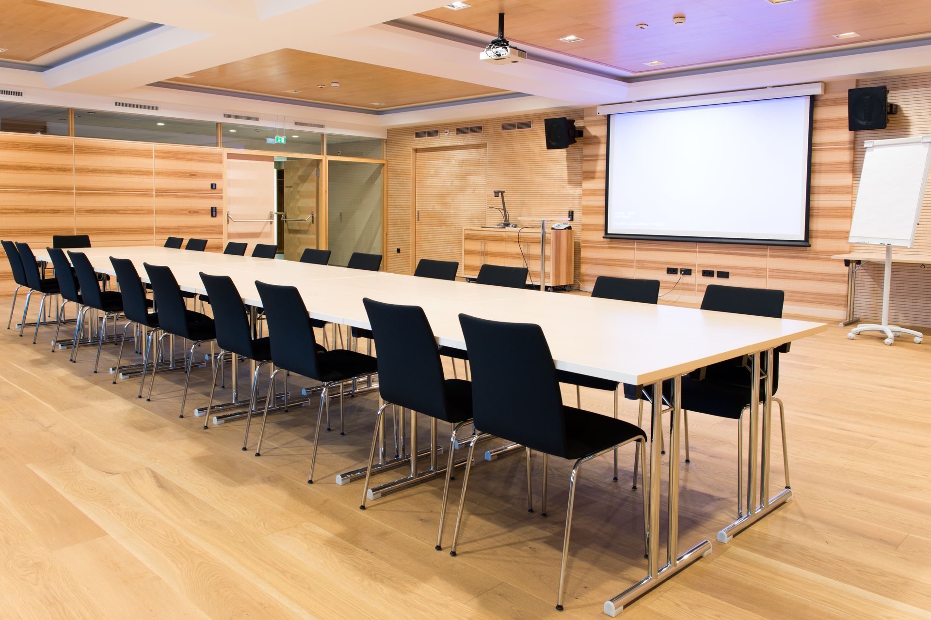 Modern conference room with long tables, black chairs, wood flooring, and a projector screen at the front