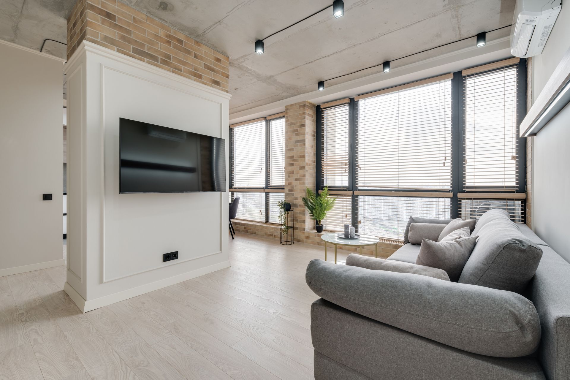 Modern bright living room with gray sofa, wall-mounted TV, large windows, and light wood flooring