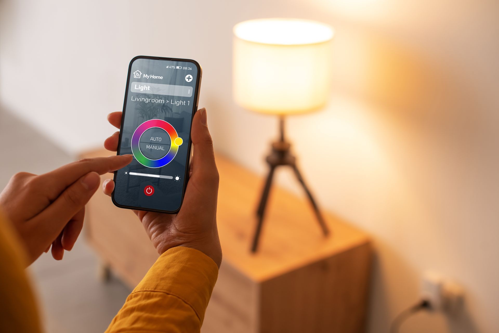 Hands holding a smartphone with a color wheel app, aimed at a lit lamp in a cozy room.