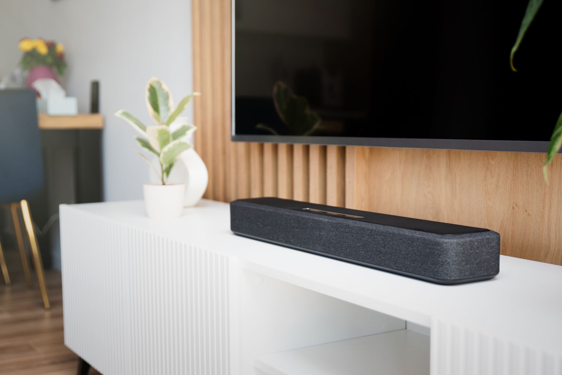 Black soundbar on a white TV console with a small potted plant beside a wall-mounted TV.