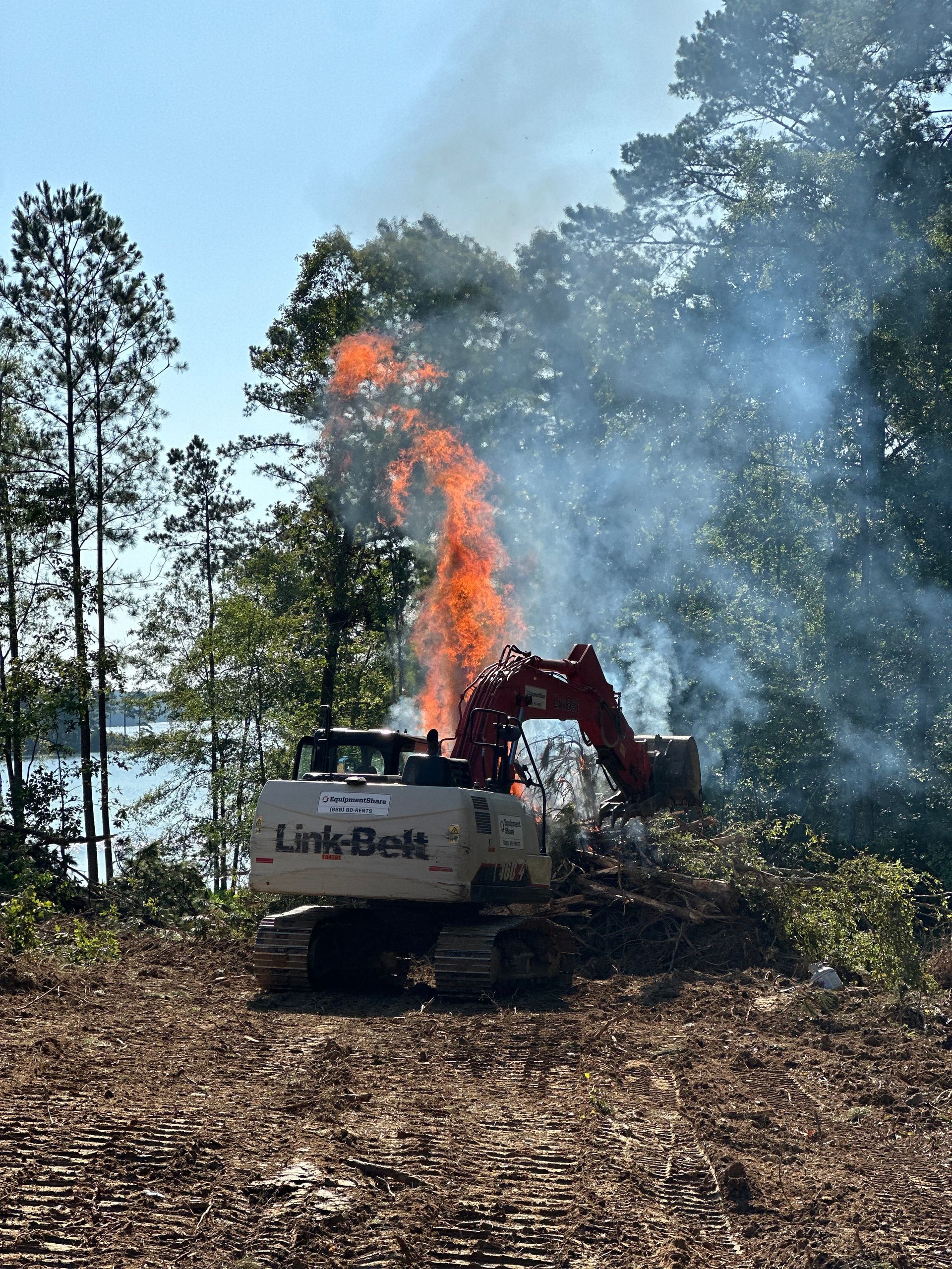 Excavator clearing brush on a hillside, trees ablaze with orange flames and smoke billowing into a clear blue sky.
