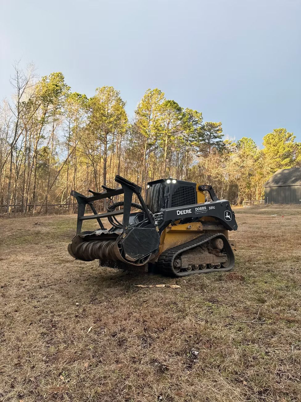 Yellow skid steer with a mulching head in a field, trees in the background.