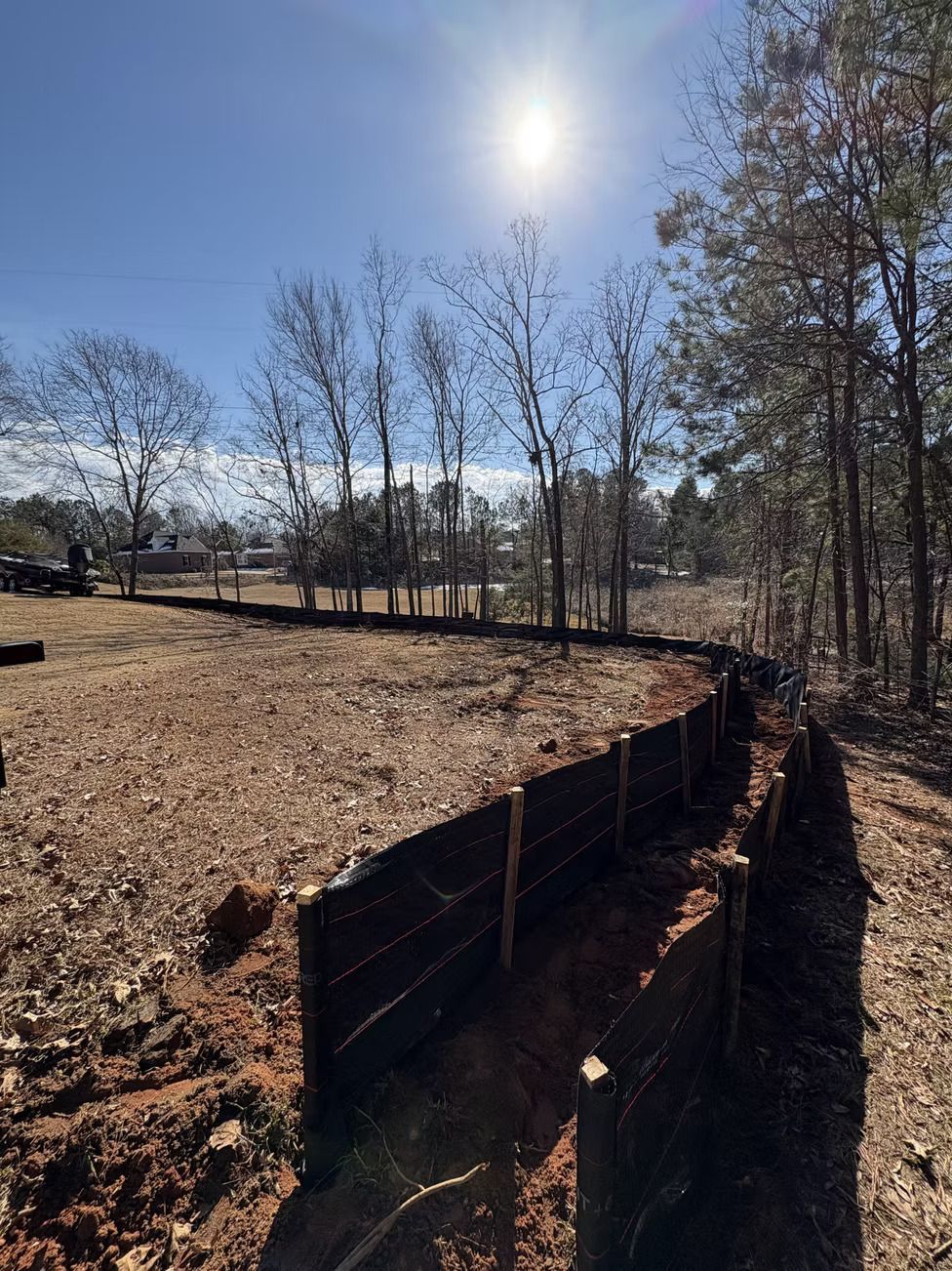 A sunny construction site with a silt fence, trees, and a dirt area.