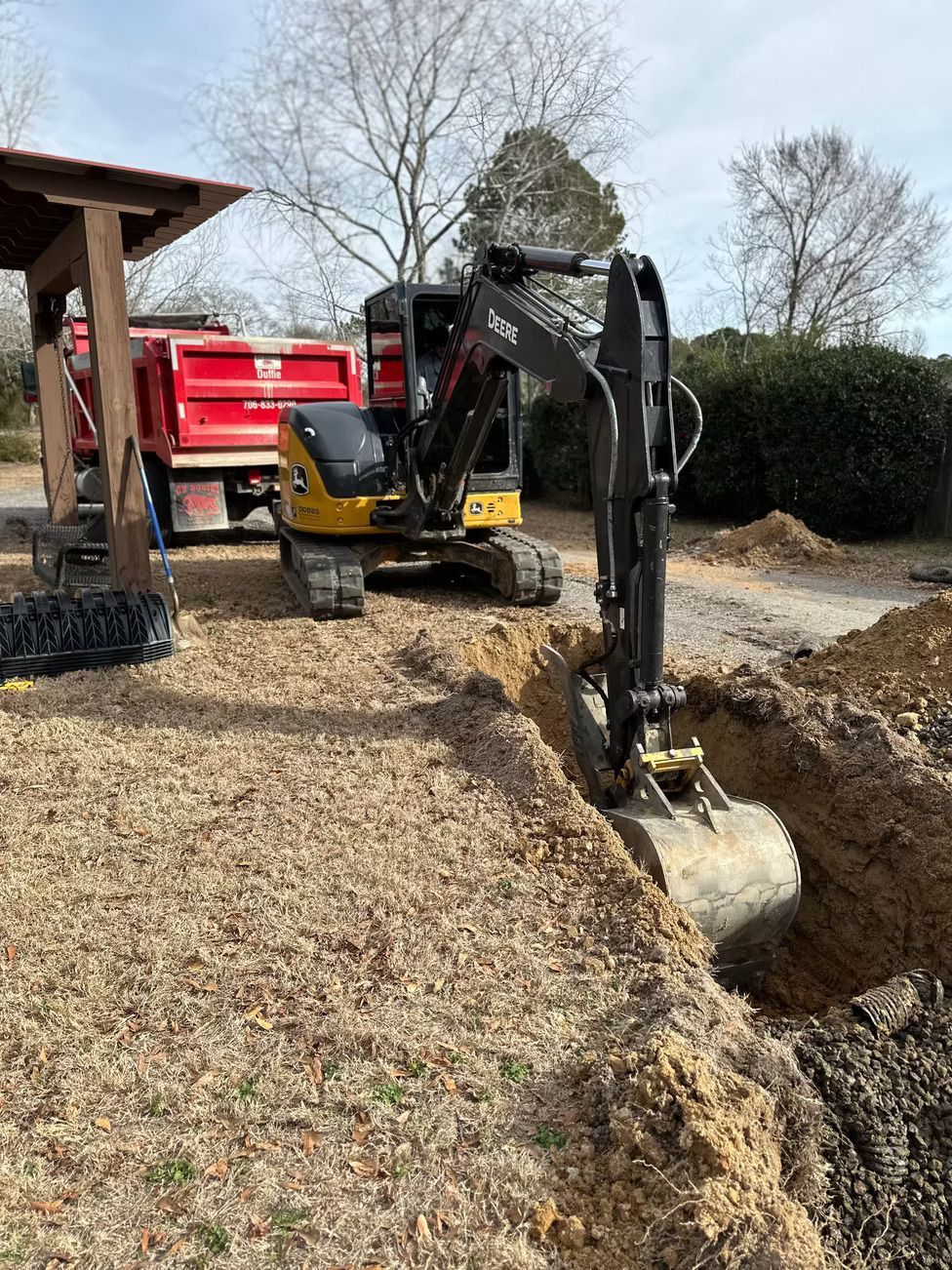 Mini excavator digging a trench on a gravel driveway, red dump truck in the background.