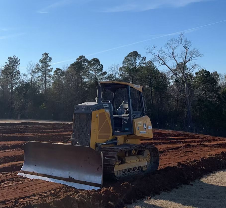 Yellow bulldozer grading red dirt in an open field, trees in the background, clear blue sky.