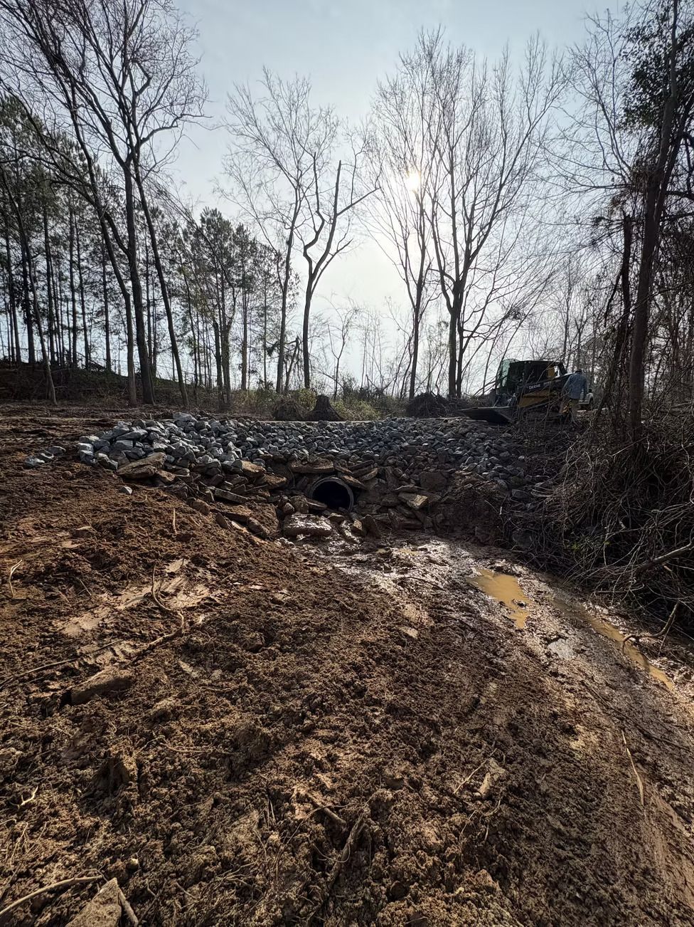 Muddy construction site with a culvert, rocks, trees, and a bulldozer under a bright sky.