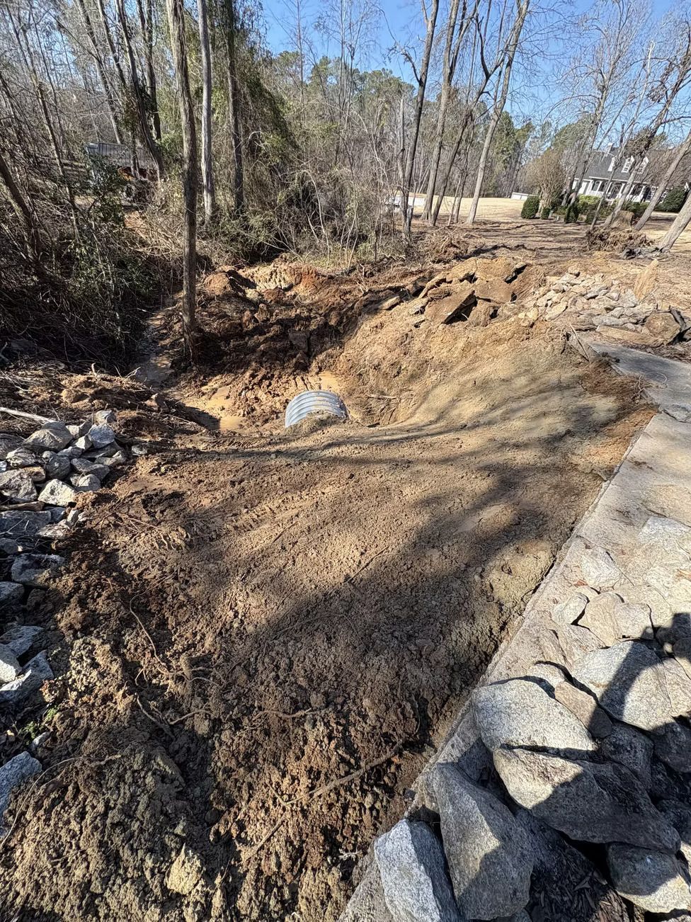 Excavated dirt bank with a culvert, lined with rocks, and trees in the background.