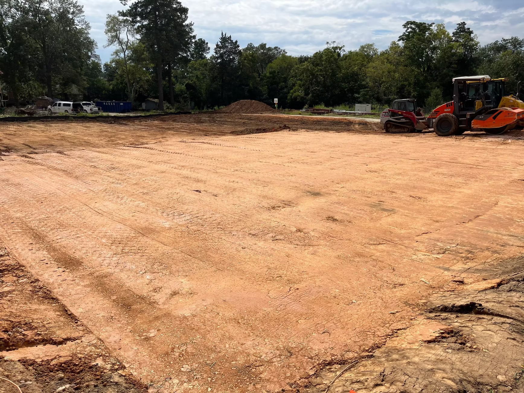 A construction site with a leveled dirt area, construction equipment, and trees in the background.