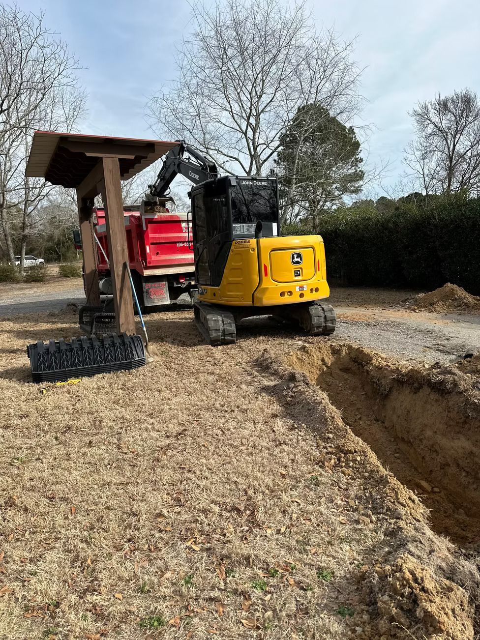Yellow excavator digging a trench next to a red dump truck. Wood structure overhead. Dirt ground with trees in the background.