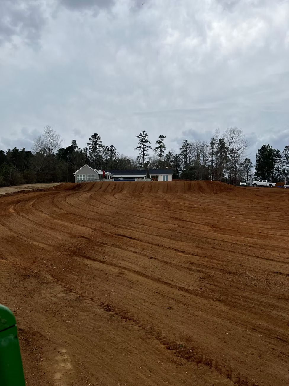 Dirt field graded, with a house on a small hill in the background under a cloudy sky.