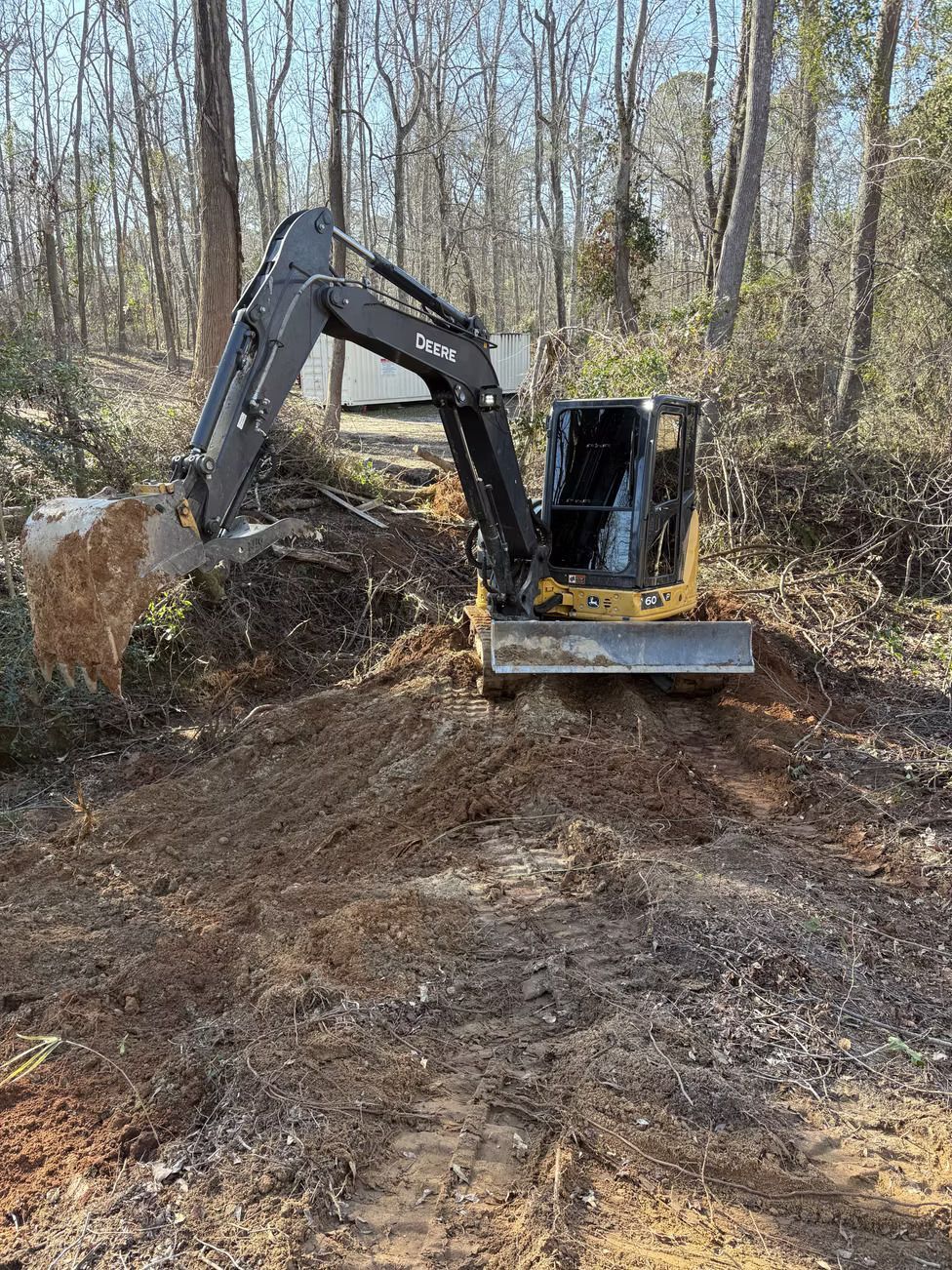 Small yellow excavator digging into a dirt pile outdoors with trees in the background.