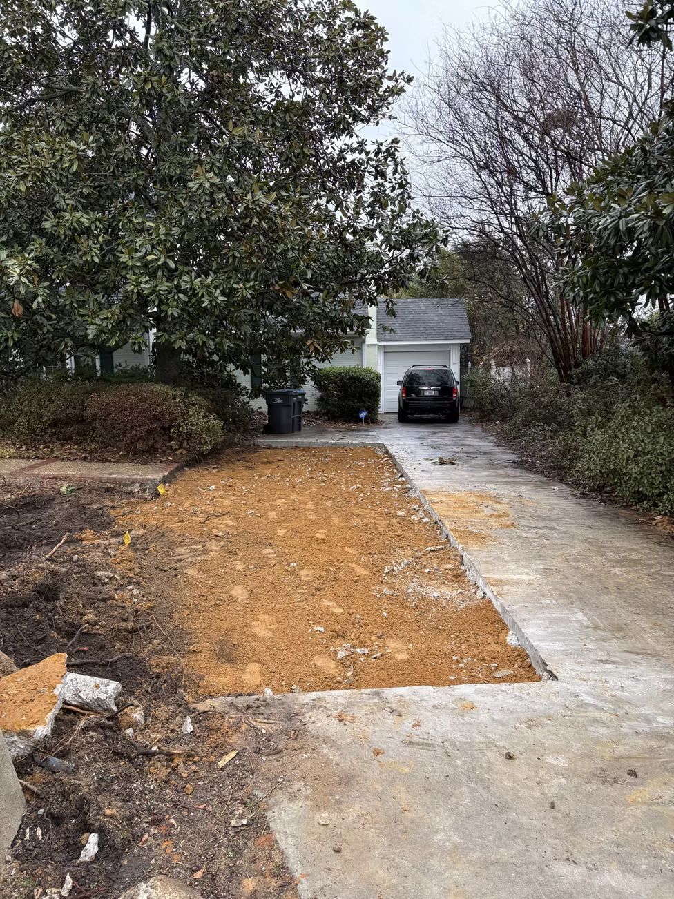 Driveway with a dirt patch, car in garage, trees.