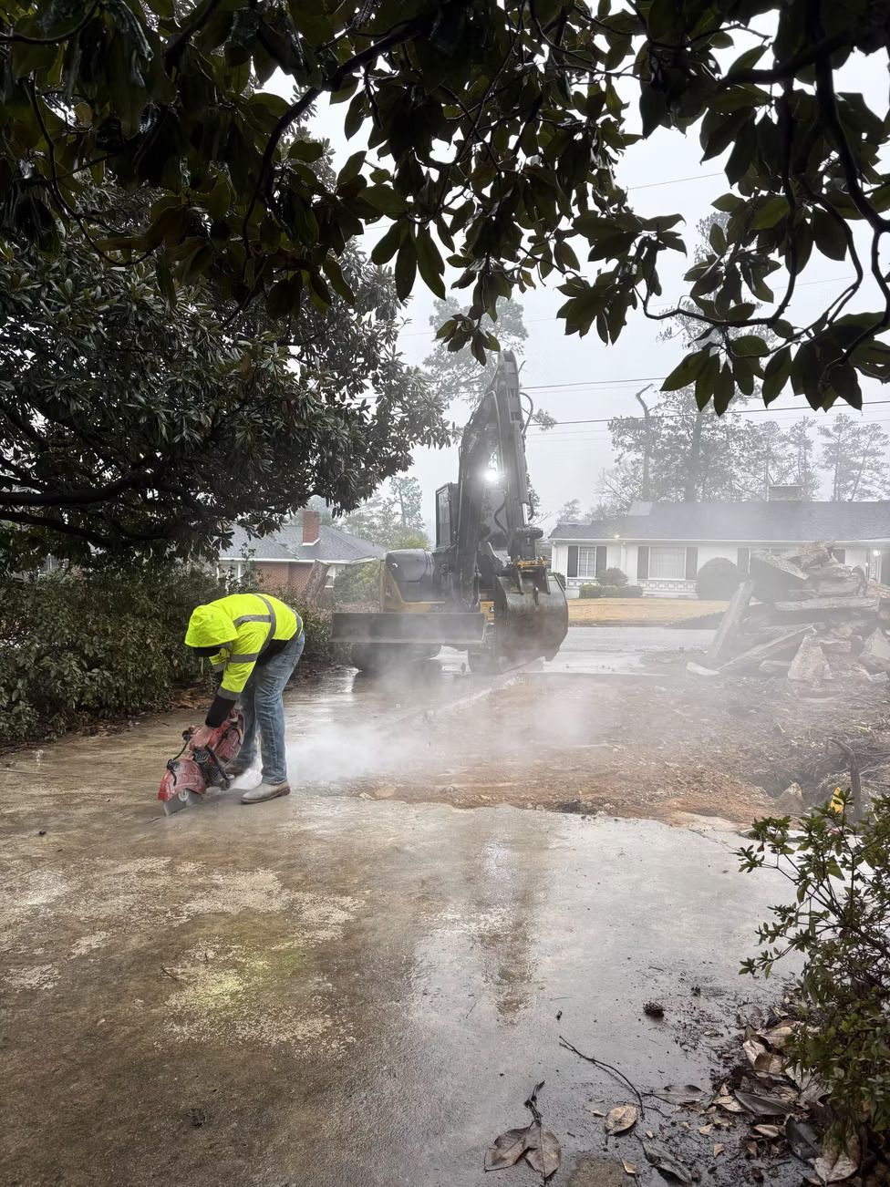 A person in safety gear cuts into pavement, creating dust. An excavator sits nearby; a rainy day setting.