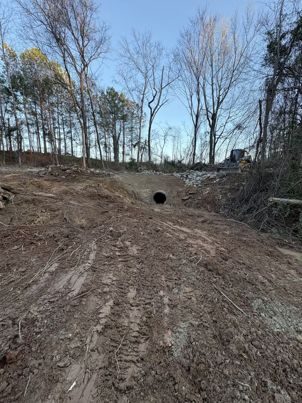 Muddy slope leading to a dark culvert under bare trees, with a hint of sky visible.