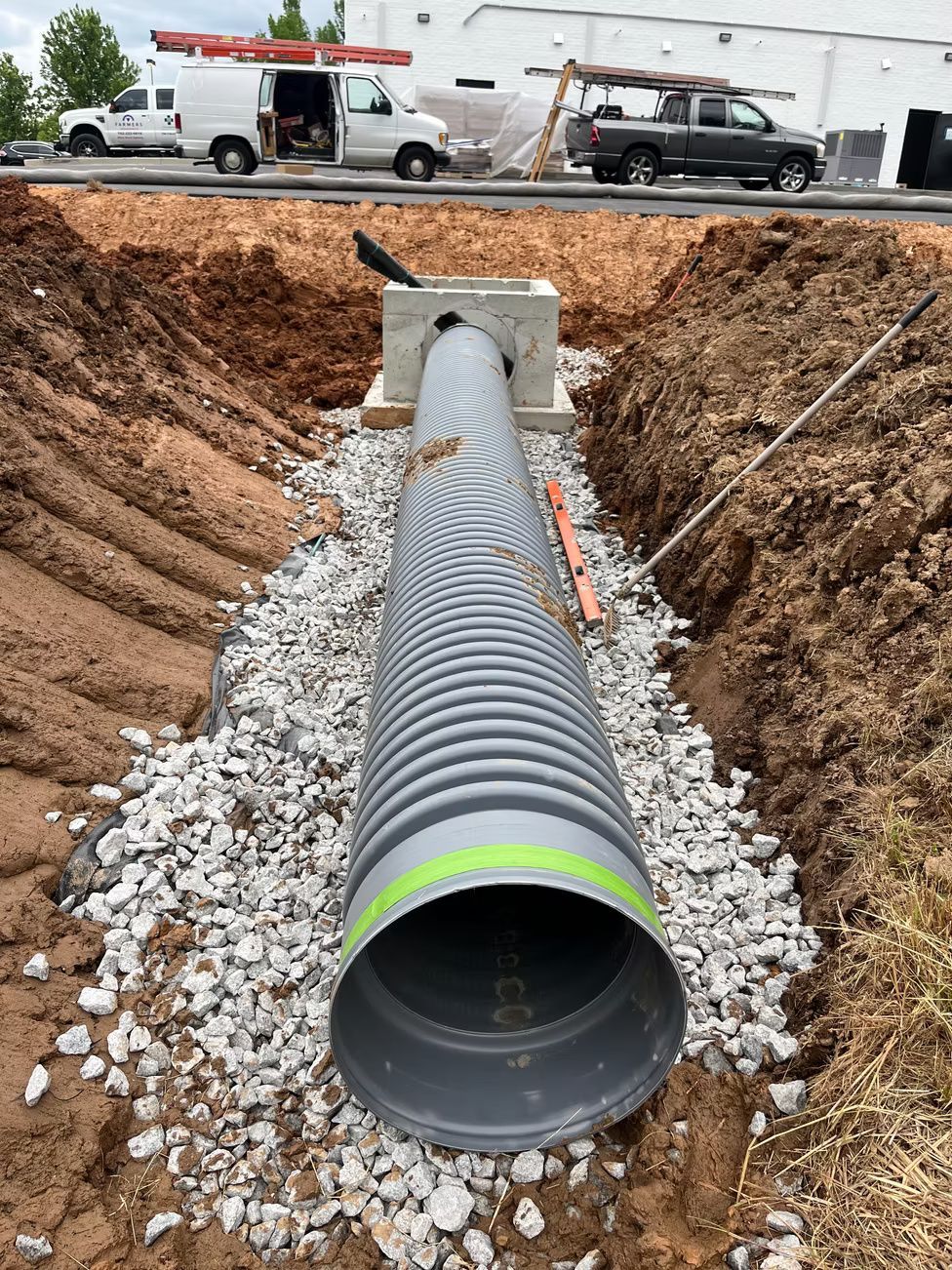 Construction site: corrugated pipe in a trench, surrounded by gravel, leading to a concrete structure, with trucks and a building in the background.