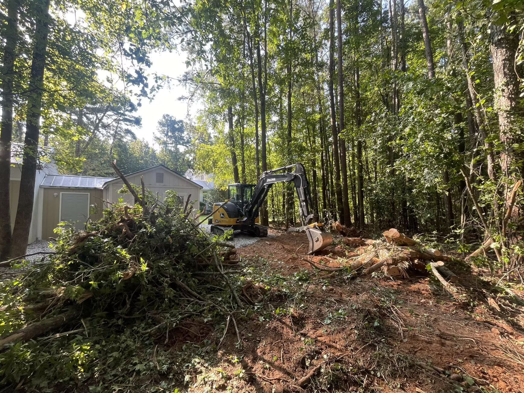 Excavator clearing land near a house surrounded by trees; logs and branches on the ground.
