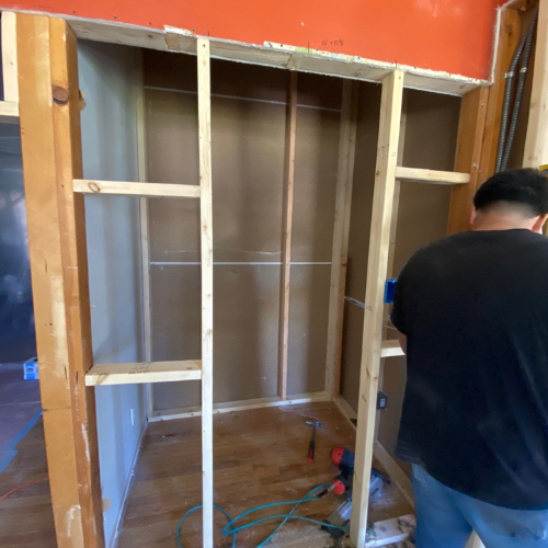 Man building wooden shelving inside a wall. Orange paint visible above.